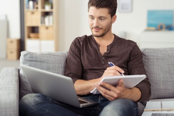 Ein junger Mann sitzt mit Laptop und Unterlagen auf dem Sofa in seiner Wohnung.