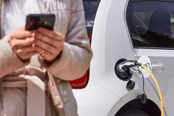 Junge Frau, die ein Elektroauto an einer öffentlichen Ladestation auflädt und mit einem Mobiltelefon bezahlt.
